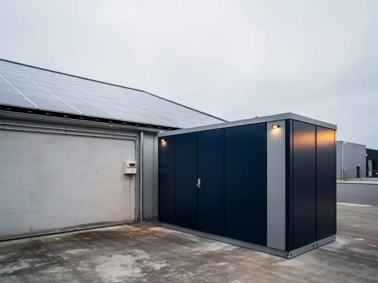 Industriële batterijopslag naast een modern magazijn met zonnepanelen op het dak en een elektriciteitsmeter aan de gevel.