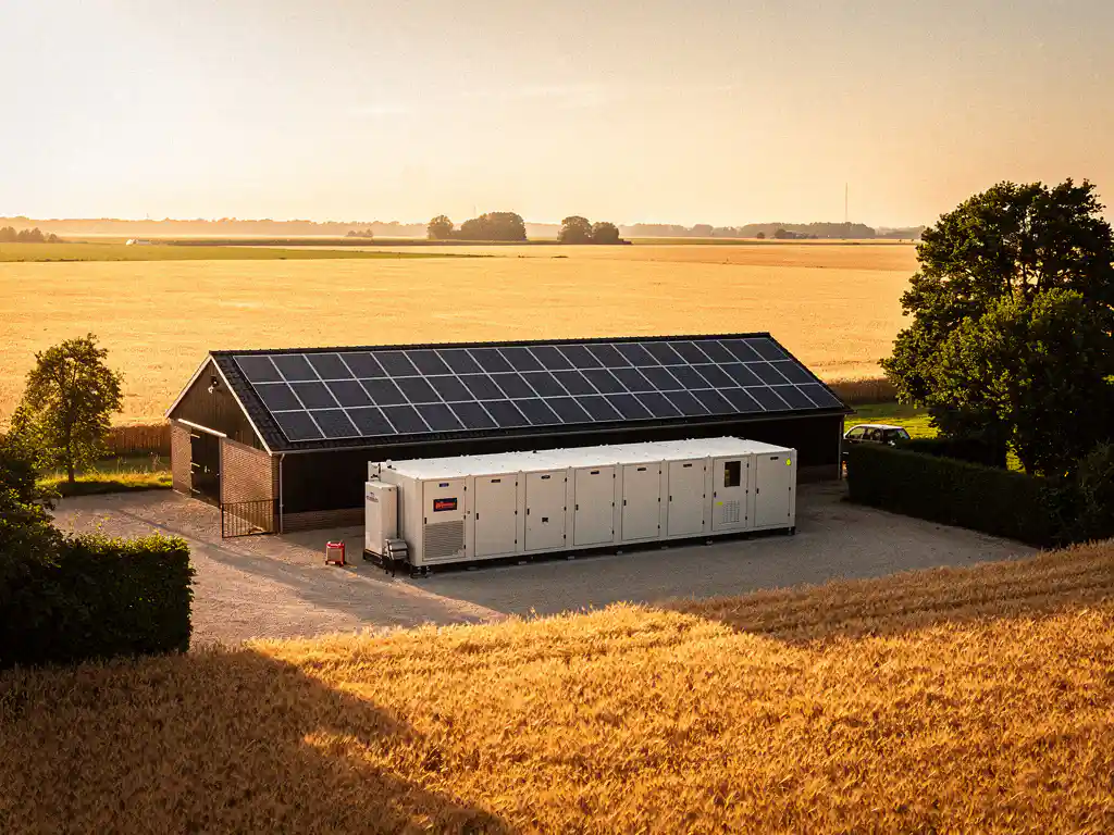 Industriële batterijopslag naast een Nederlandse schuur met zonnepanelen, omgeven door gouden tarwevelden in het late middaglicht.